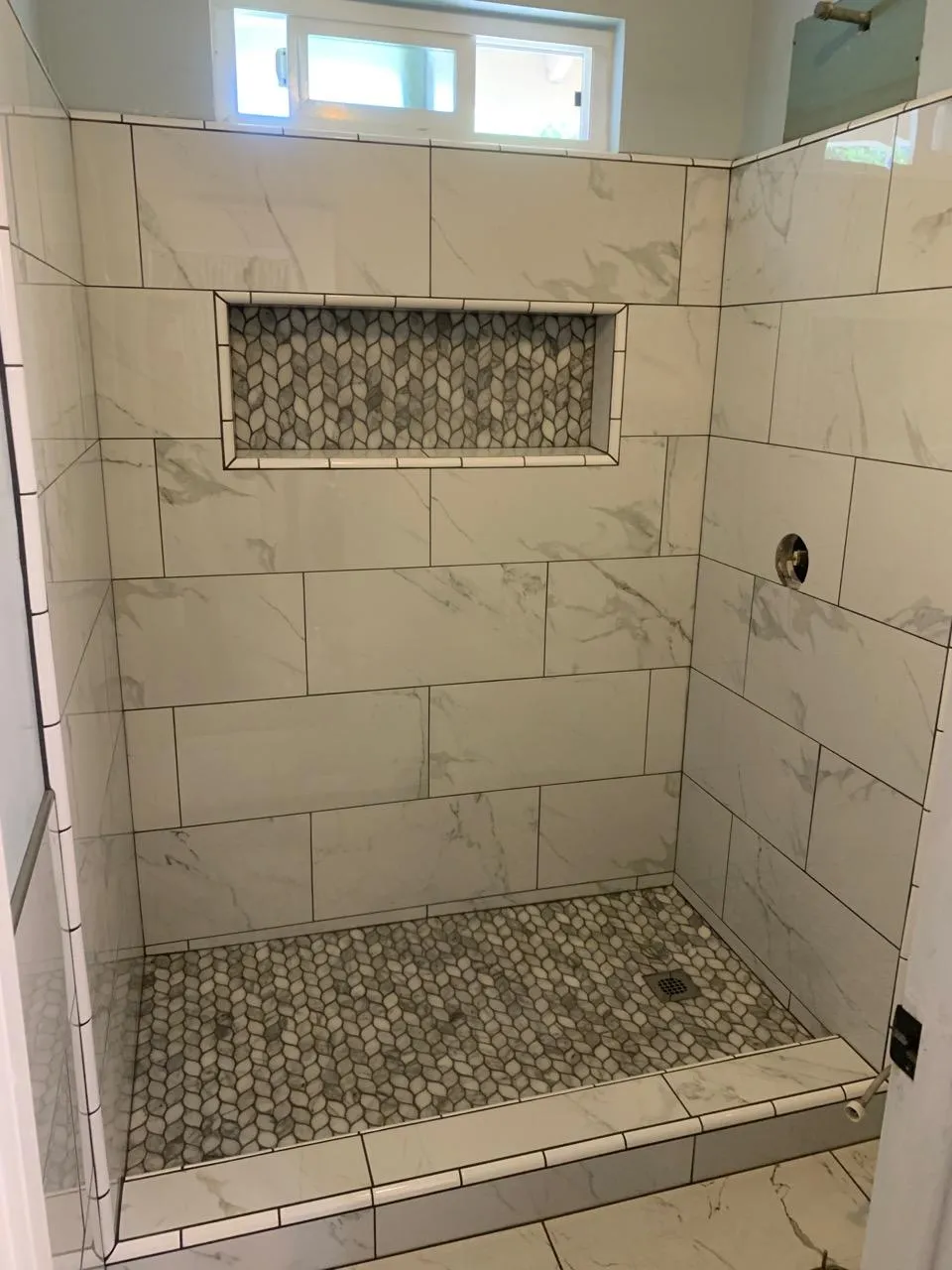 Walk-in shower with light stone wall tiles, pebble mosaic floor, and recessed mosaic niche