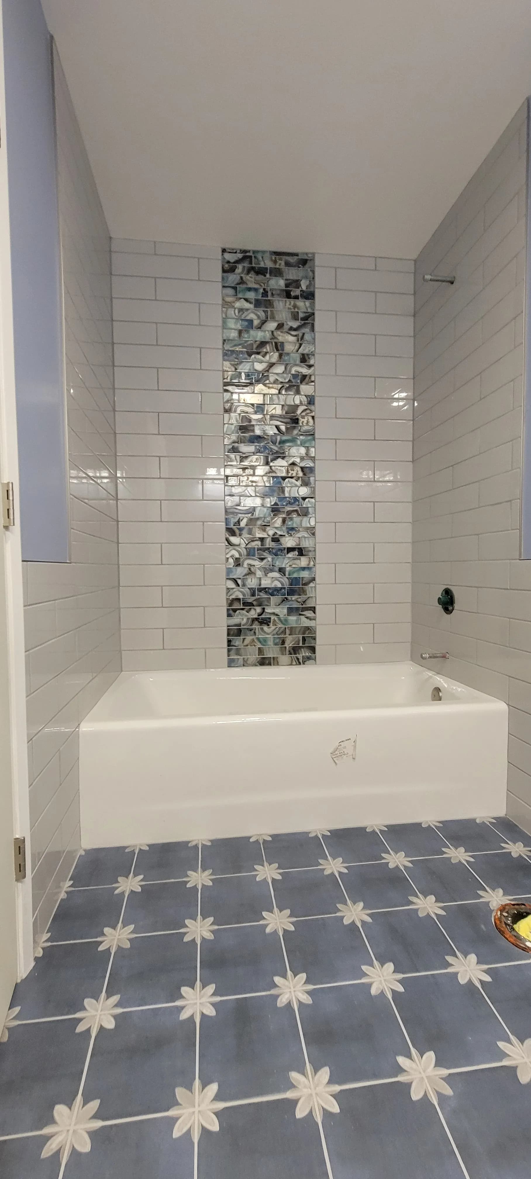 Bathtub shower with white tile walls, vertical mosaic accent strip, and patterned tile floor